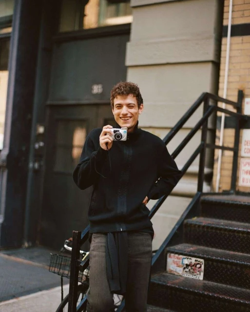 Young man with curly hair stands outside holding a digital camera, smiling on stairs next to a brick building with a metal staircase.