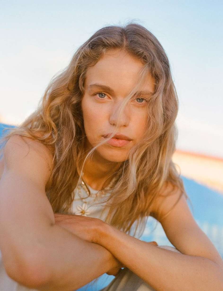 Close-up portrait of a woman with long blonde hair, blue eyes, and arms crossed in front of a light blue and sandy-beige backdrop.