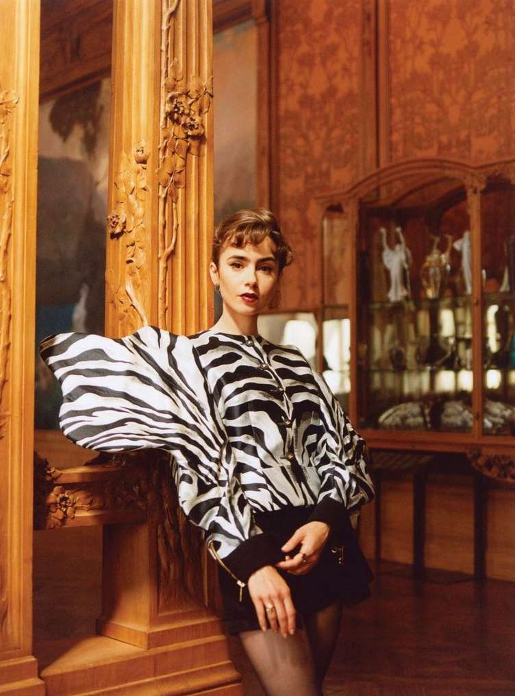 Elegant woman in zebra-print blouse leans against carved wooden column indoors. Fashionable pose with dark hair, neutral face and sophisticated style.