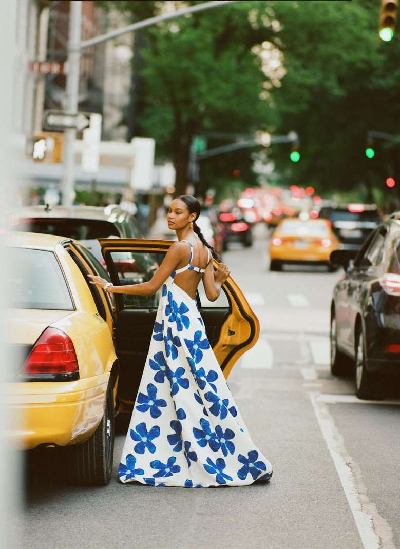 Adult female exiting a yellow taxi cab in an urban setting. She is wearing a white dress with blue floral pattern. Cars and buildings visible.