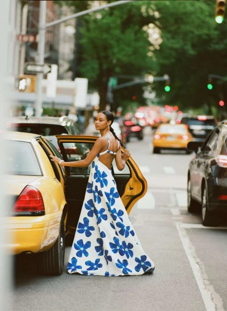 Adult female exiting a yellow taxi cab in an urban setting. She is wearing a white dress with blue floral pattern. Cars and buildings visible.
