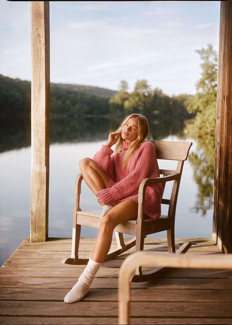 A blonde woman wearing a pink sweater and white socks sits in a wooden chair on a wooden dock in front of a lake and green trees.