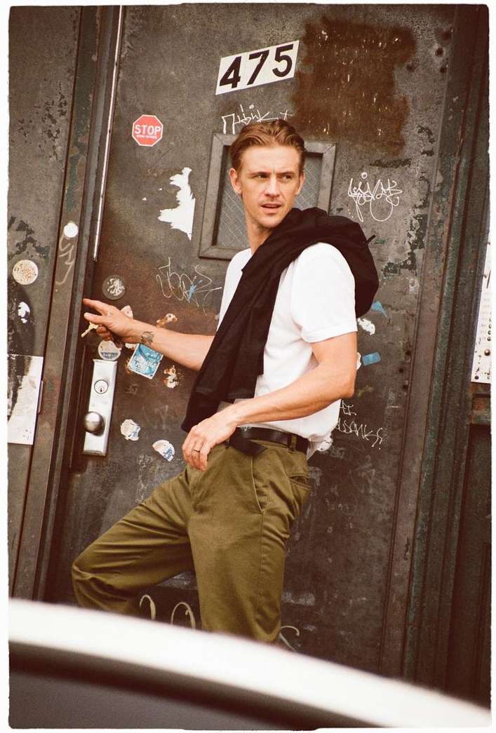 Adult male in a white t-shirt and olive pants poses against an old, graffitied door with a dark jacket slung over his shoulder.