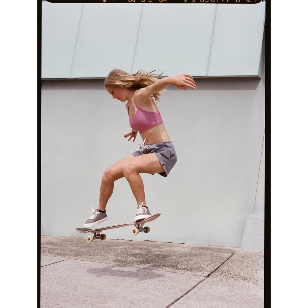 An athletic young woman in sportswear airborne on a skateboard against a neutral wall backdrop, showcasing urban sport and athleticism.