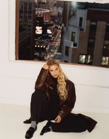 Blonde woman seated on the floor wearing a brown leather jacket and loafers against a white wall with a city window view