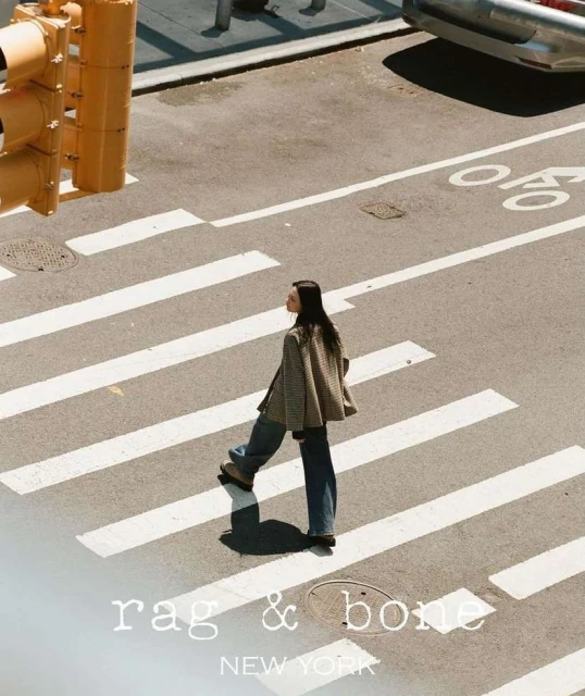 An adult woman in casual clothing confidently strides across a zebra-striped crosswalk in an urban setting. A traffic light is visible.