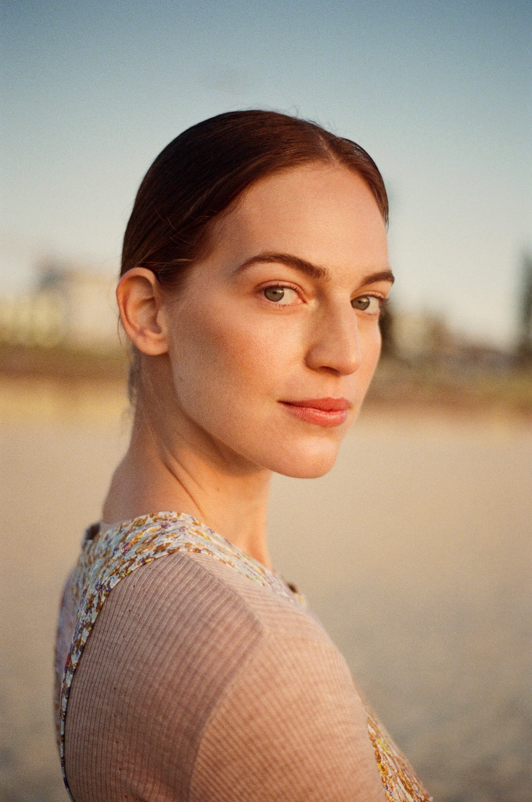Portrait of a woman with brown hair and a pink patterned top outdoors on a sandy beach during golden hour.