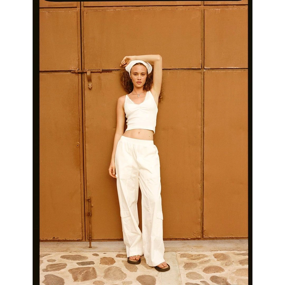 Woman in white tank top and white trousers posing against a brown metal door with her arm raised behind her head
