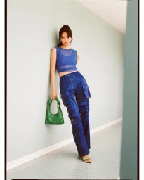 Woman in blue mesh crop top and shiny blue cargo trousers leaning against a pale turquoise wall holding a green handbag