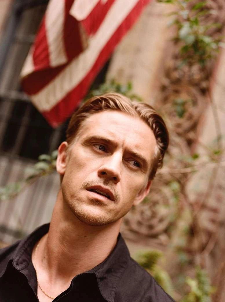 Portrait of a man in a dark shirt looking away with an American flag hanging in the background by Ryan Brabazon