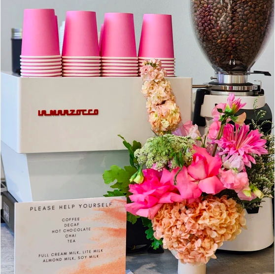 Coffee preparation setup with a La Marzocco espresso machine, pink cups, a grinder with coffee beans, and a flower arrangement.