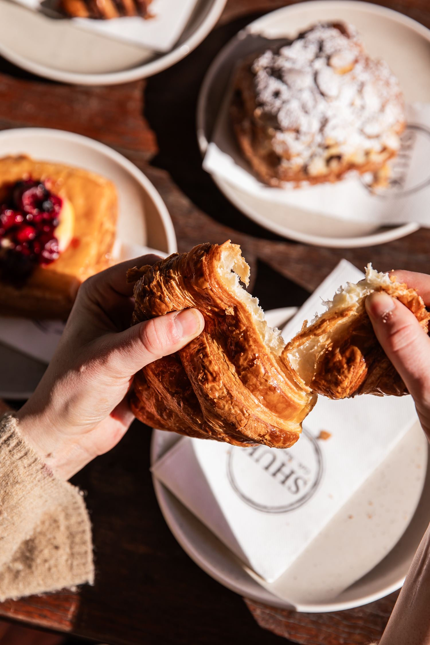Overhead shot of hands tearing a croissant, revealing its flaky layers, with plates of pastries on a wooden table in soft daylight.