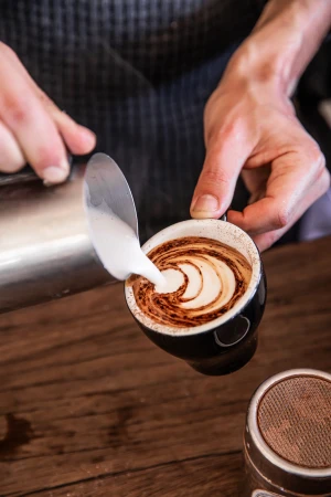 Barista pours foamed milk into espresso cup, creating latte art on a wooden counter, with a focus on the drink and coffee preparation.