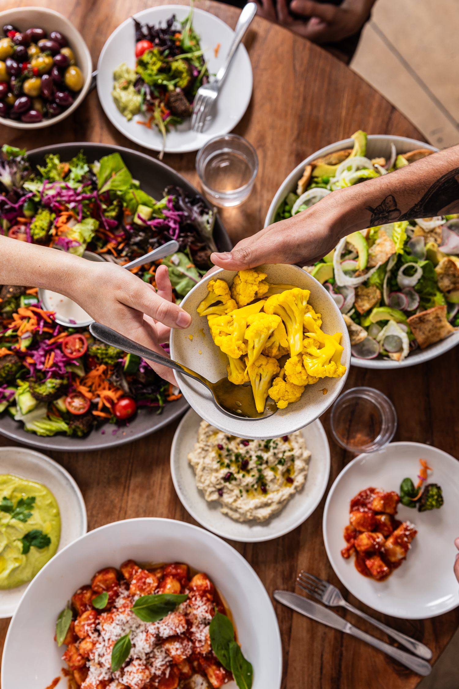 Overhead shot of a wooden table laden with a variety of vibrant dishes including colorful salads, gnocchi, cauliflower, and dips.