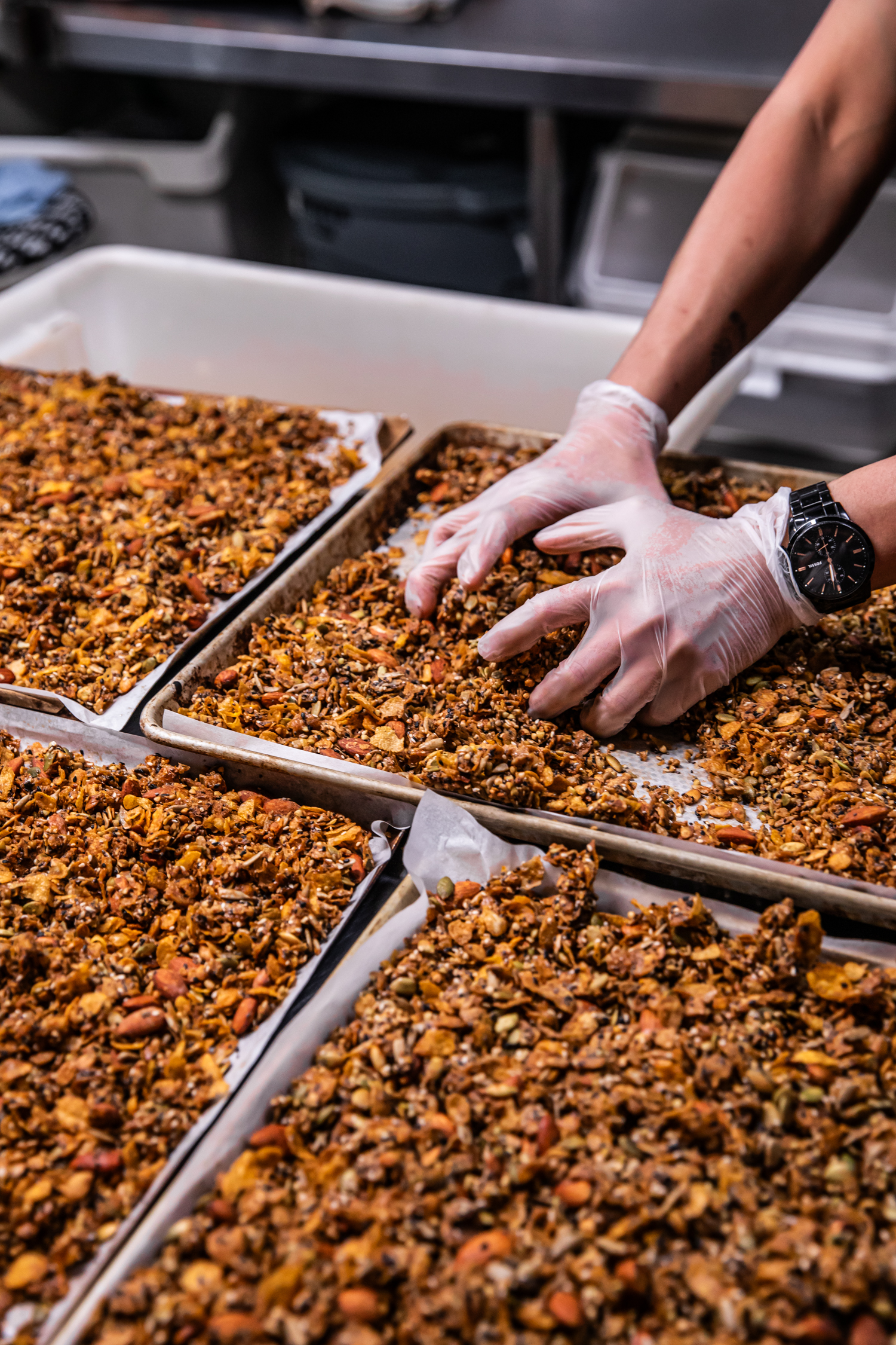Close-up shot of gloved hands working granola on baking trays in a commercial kitchen, showcasing food preparation and artisanal food production.