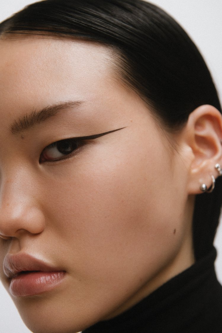 Close-up portrait of an East Asian woman with graphic winged eyeliner and silver ear piercings wearing a black top.