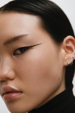 Close-up portrait of an East Asian woman with graphic winged eyeliner and silver ear piercings wearing a black top.