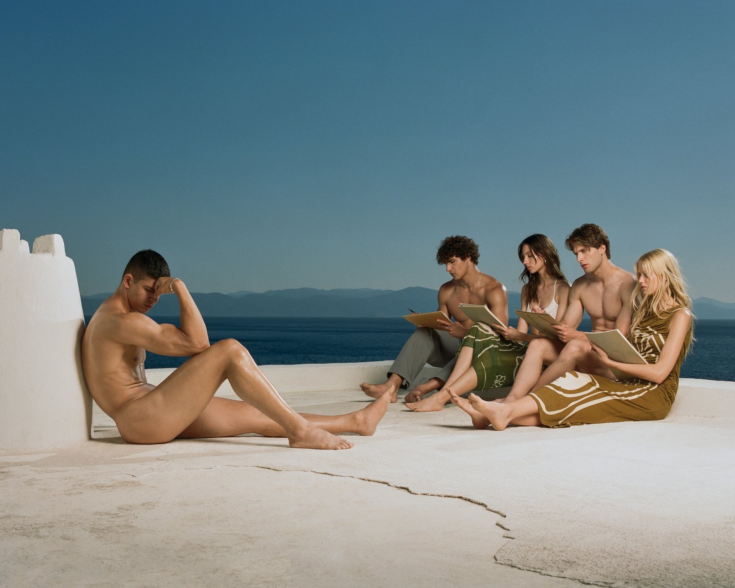 Nude male model poses outdoors as artists sketch him; blue sea and sky backdrop. Live figure drawing in bright daylight.