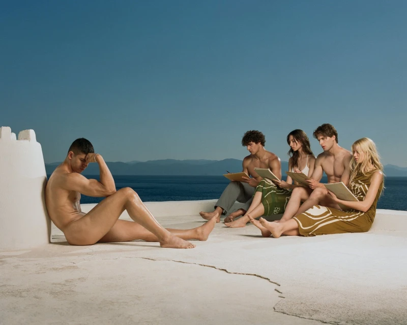 Nude male model poses outdoors as artists sketch him; blue sea and sky backdrop. Live figure drawing in bright daylight.