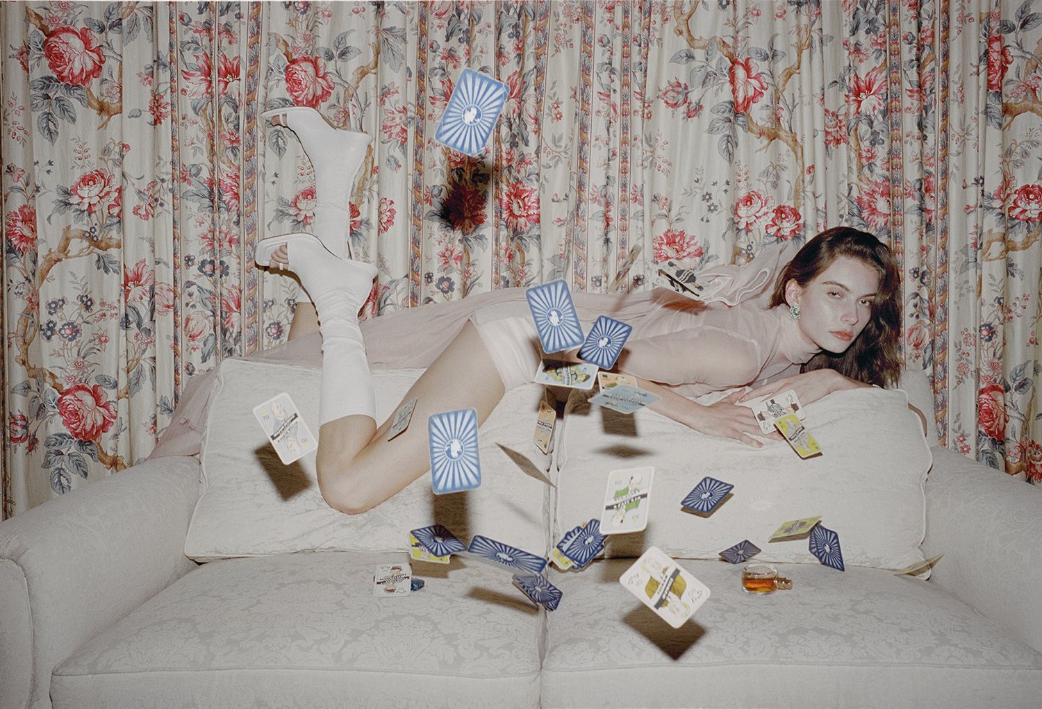Adult female poses on a couch surrounded by playing cards. She wears white boots and the room features a floral background.