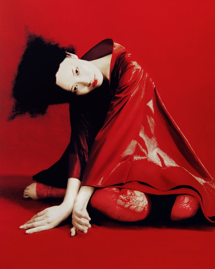 Asian woman in red kimono-style dress seated against a red backdrop in studio shot. Dark hair contrasts with pale skin and bright colours.