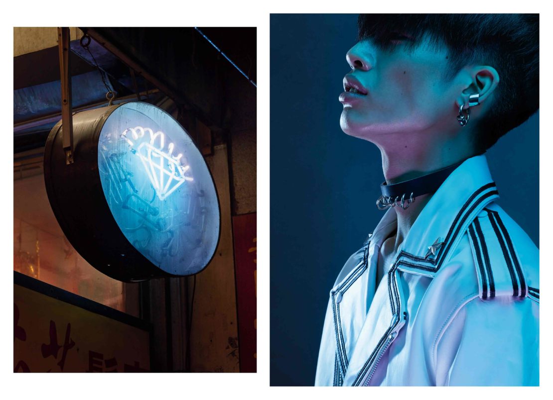 Close-up shot of a person with lip piercings wearing a choker and jacket next to a neon diamond light in an urban environment.