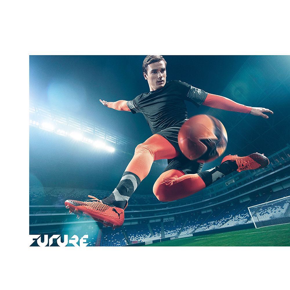 A male soccer player wearing Puma sportswear in mid-air kicking a soccer ball in a stadium filled with fans, orange and black shoes.