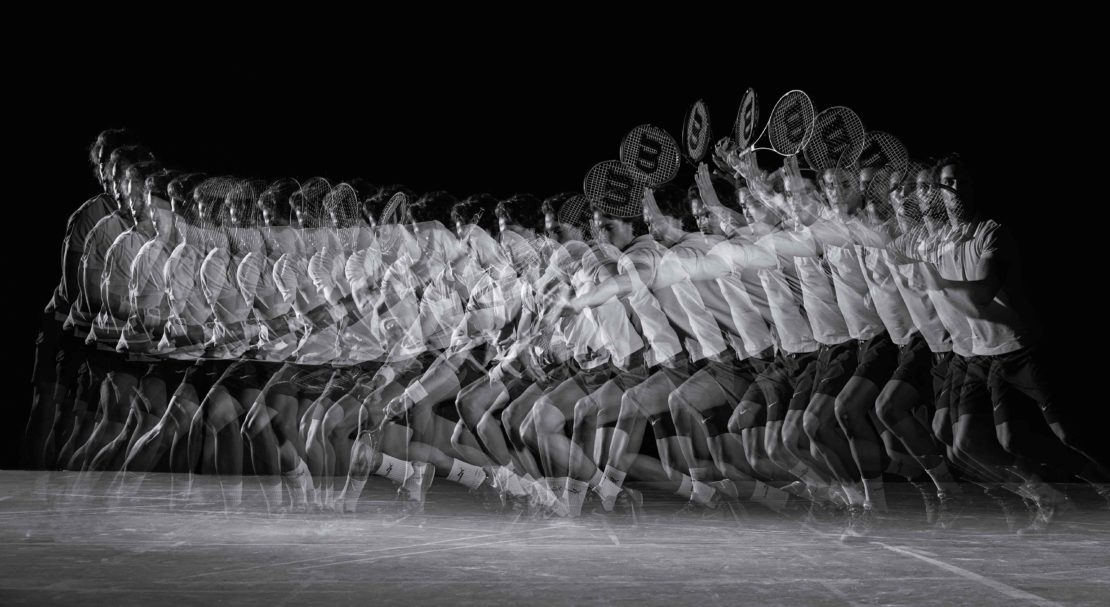 Monochrome stroboscopic image of a man in motion hitting a tennis ball with a racquet, showcasing movement against a black background.