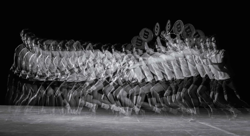 Monochrome stroboscopic image of a man in motion hitting a tennis ball with a racquet, showcasing movement against a black background.