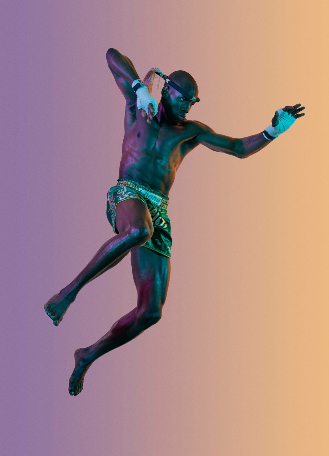 Muscular man in Thai boxing stance mid-air, wearing traditional shorts, hand wraps, and headgear, against a gradient background.