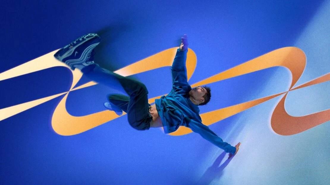 Man in mid-air wearing a blue jacket and sneakers against a blue background with orange swirl patterns, captured in a studio shot.