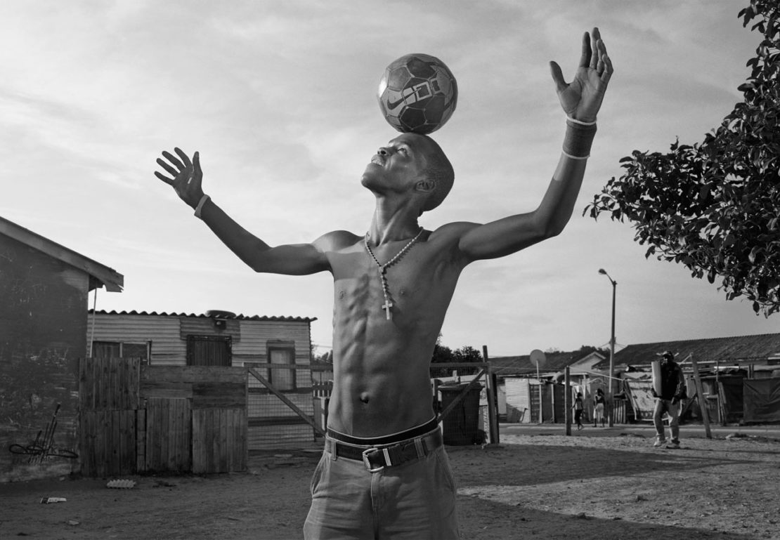 Monochrome portrait of shirtless man balancing soccer ball on head with arms raised in an outdoor setting. Street and buildings behind him.
