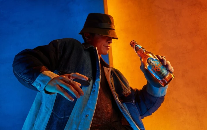 Man in bucket hat and denim jacket holding a Tiger beer bottle against a blue and orange lit background