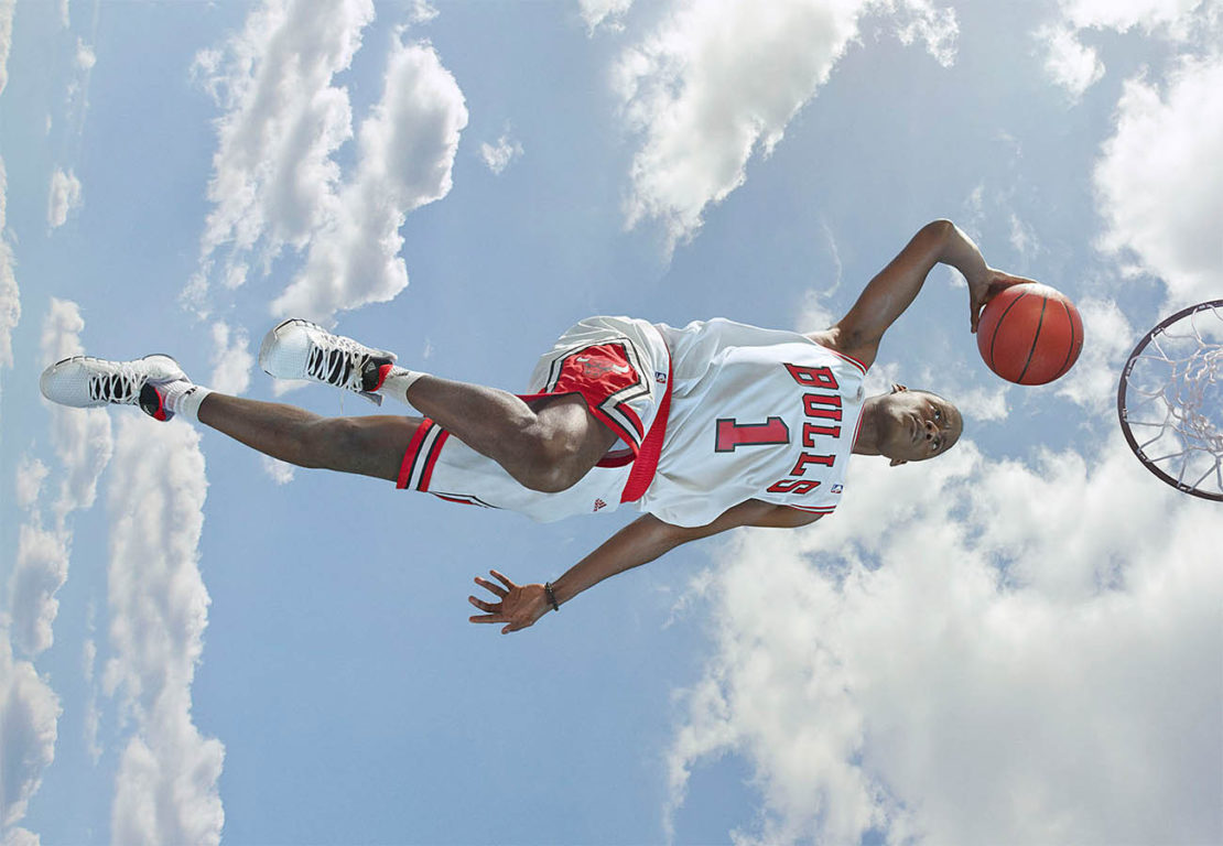 A basketball player in a Bulls jersey leaps toward a hoop, ball in hand, set against a bright sky with fluffy clouds.