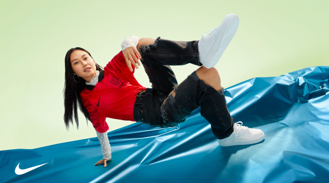 A person in a red soccer jersey and ripped jeans balances on a shiny blue surface in white sneakers against a pale green backdrop.