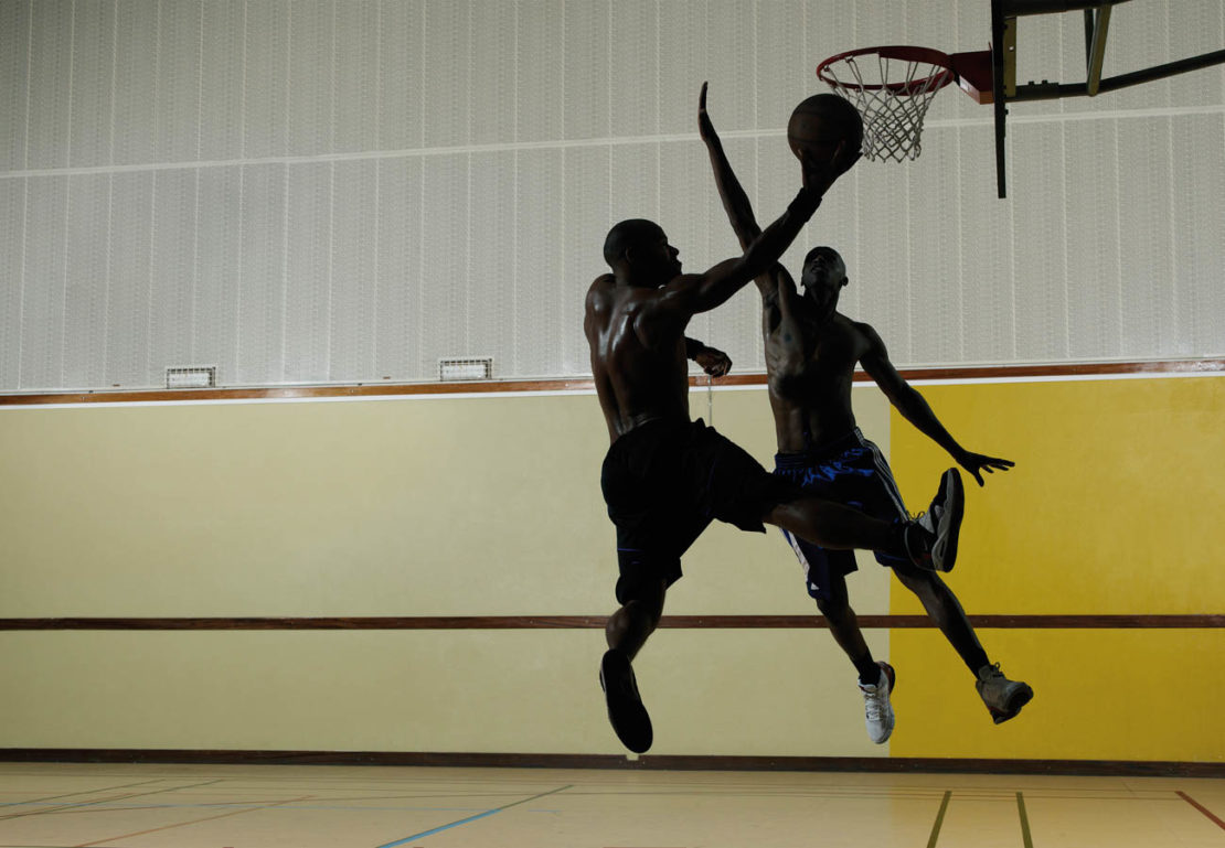 Two adult males in silhouette play basketball on an indoor court, one leaping with the ball, the other defending near the hoop.