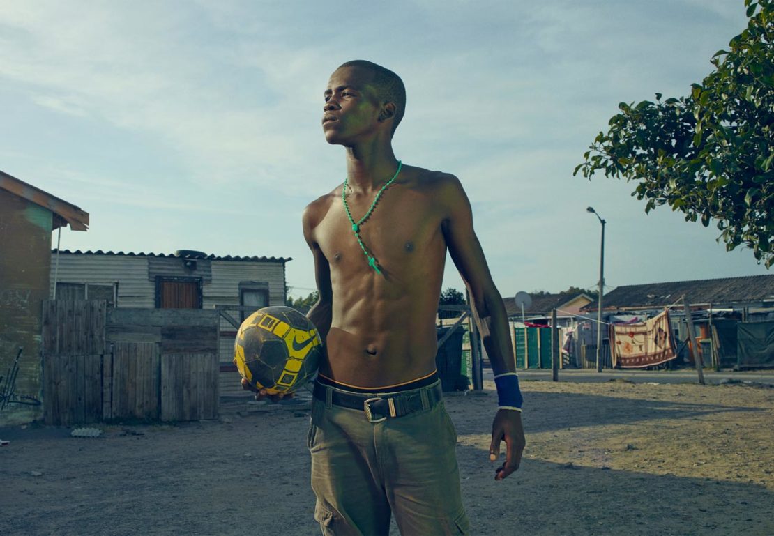 Young man stands with a soccer ball in a neighborhood, wearing a necklace, wristband, and casual pants, looking upwards in daylight.