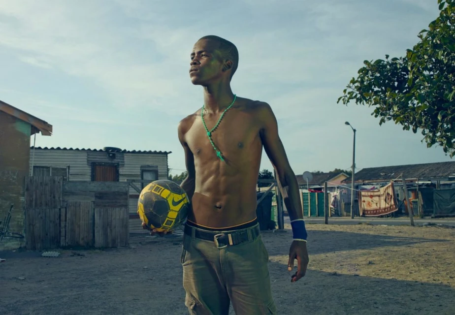 Young man stands with a soccer ball in a neighborhood, wearing a necklace, wristband, and casual pants, looking upwards in daylight.