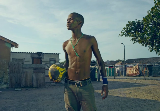 Young man stands with a soccer ball in a neighborhood, wearing a necklace, wristband, and casual pants, looking upwards in daylight.