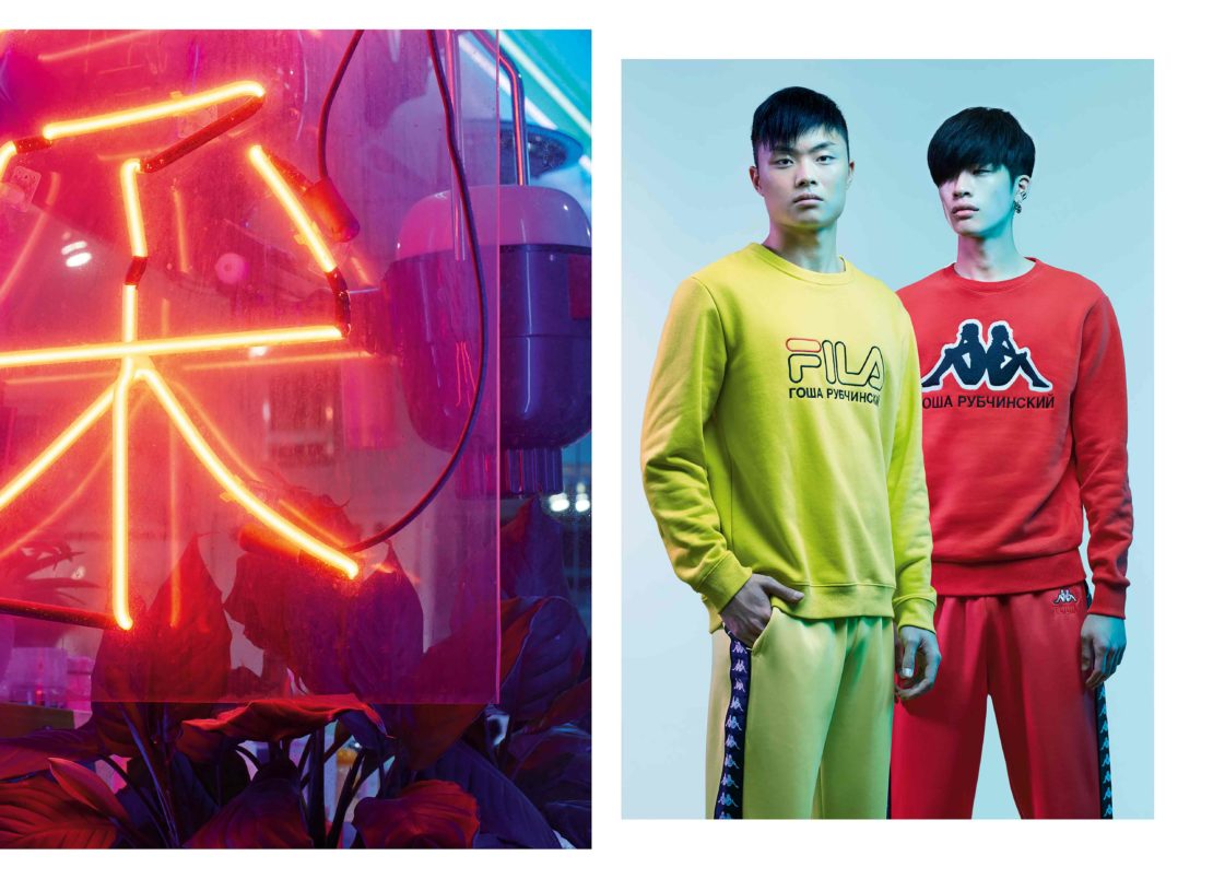 Two Asian male models wearing branded sweatshirts and matching sweatpants stand side-by-side in a studio with neon lighting.