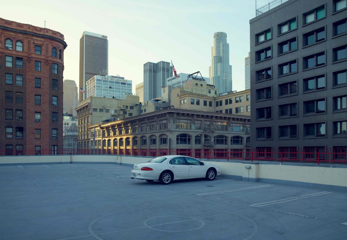 A white car is parked on the roof of a building in an urban city with skyscrapers in the background during daylight hours.