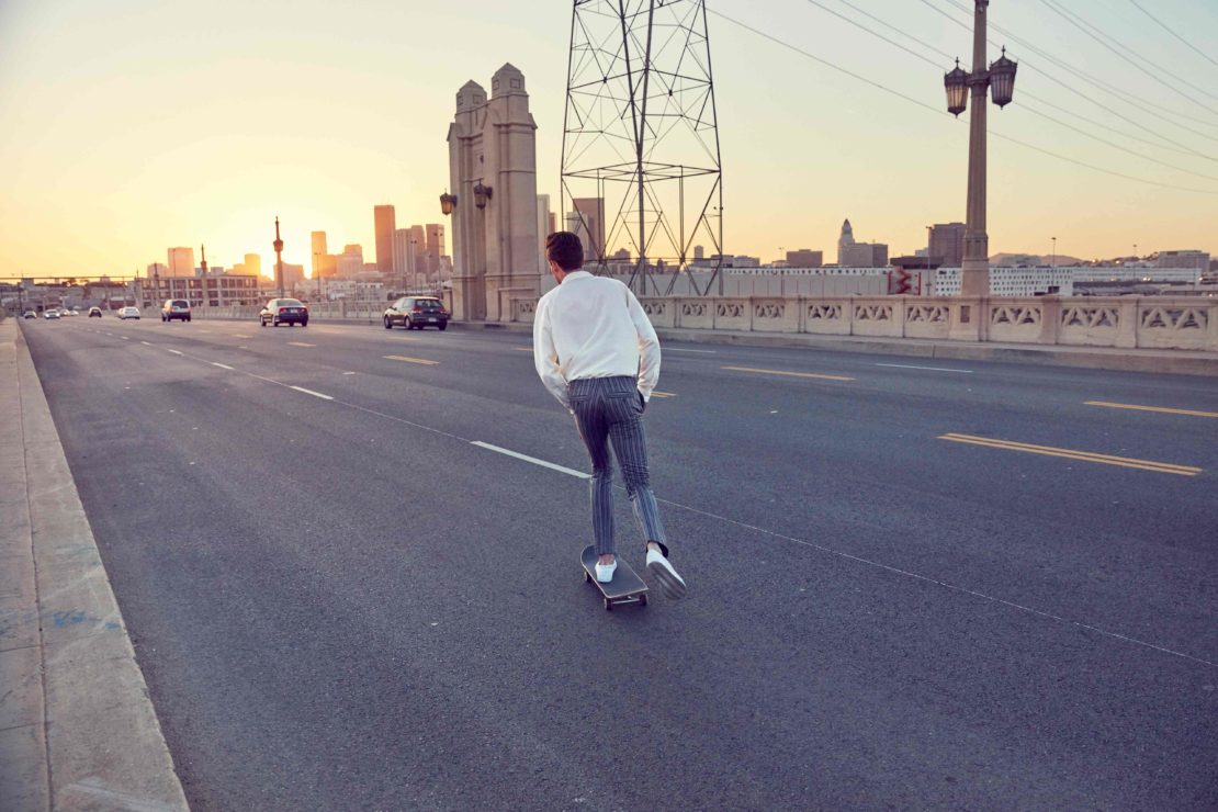 Rear view of a young adult male riding a skateboard down a city street at sunset with a cityscape silhouette on the horizon.