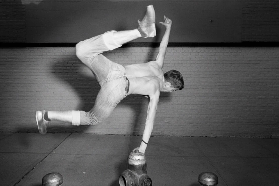 Shirtless man balances on a fire hydrant in a handstand pose against a brick wall in an urban setting in black and white.