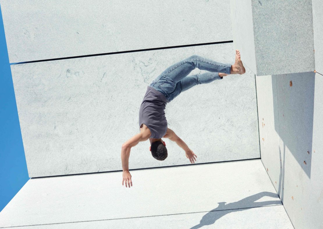 An adult in a gray tank top and blue jeans performing a handstand on a white tiled surface against a bright blue sky in an outdoor setting.