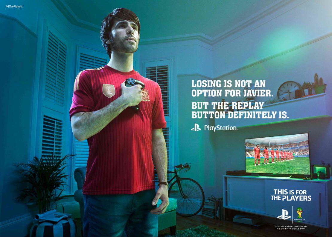 Adult male in soccer jersey holds a video game controller indoors, with television screen and bicycle visible in the background.