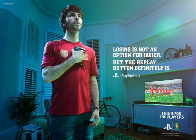 Adult male in soccer jersey holds a video game controller indoors, with television screen and bicycle visible in the background.
