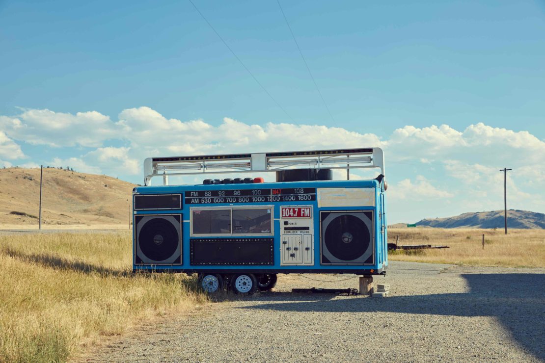Oversized blue boombox on wheels in a rural setting with open field and blue sky with clouds, novelty art installation.