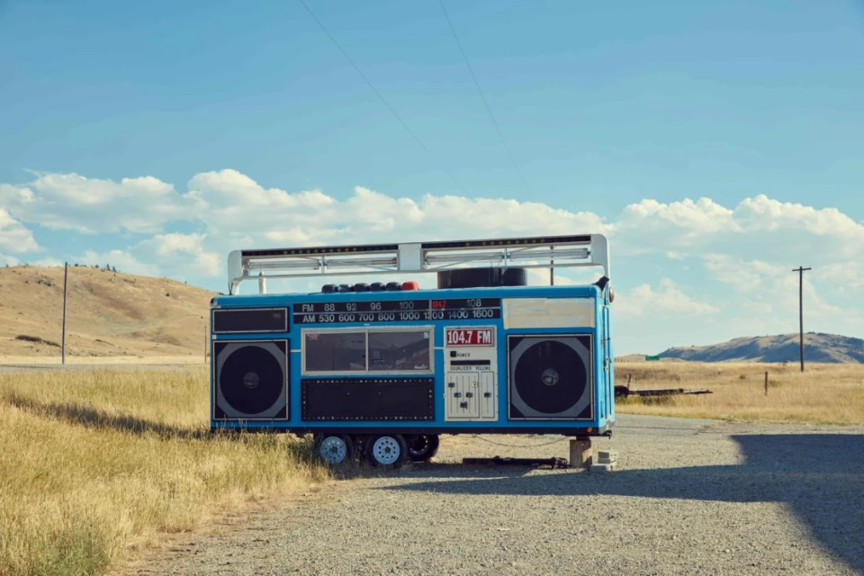 Oversized blue boombox on wheels in a rural setting with open field and blue sky with clouds, novelty art installation.