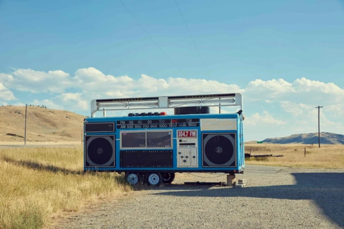 Oversized blue boombox on wheels in a rural setting with open field and blue sky with clouds, novelty art installation.