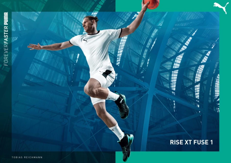 Male handball player in mid-air throwing orange ball, Puma athletic wear, indoor arena setting with teal and white structural background.